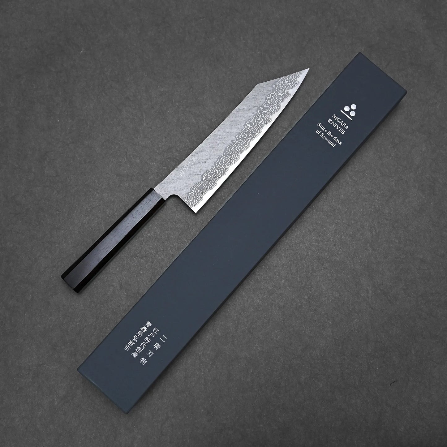 The Nigara tsuchime damascus VG10 kiritsuke gyuto 210mm, with a black handle and textured blade, is displayed beside a rectangular black box with Japanese and English writing on a dark background.