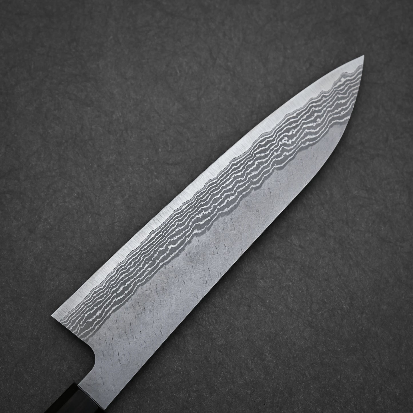 A close-up of the Nigara Hamono Nigara tsuchime damascus VG10 gyuto 210mm, showcasing its wavy Damascus steel blade pattern, rests on a dark textured surface.