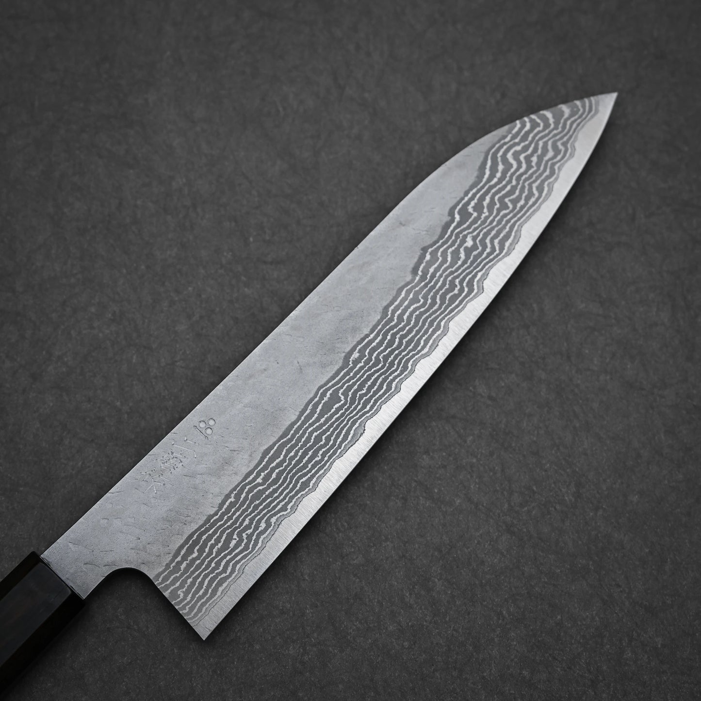 A close-up of the Nigara Hamono Nigara tsuchime damascus VG10 gyuto 210mm knife, showcasing its wavy-patterned blade on a dark textured background.