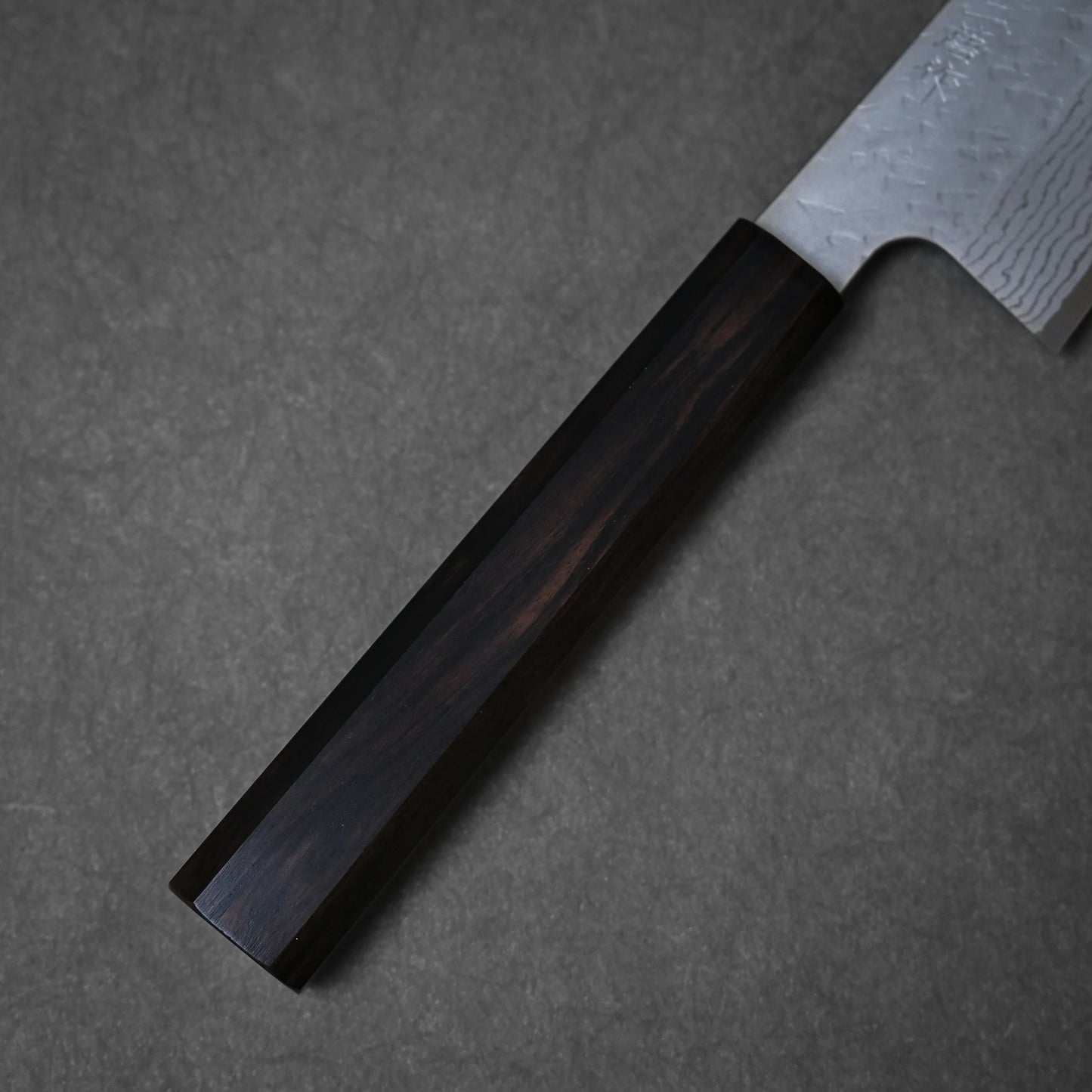 A close-up of the Nigara Hamono Nigara tsuchime damascus VG10 gyuto 210mm shows its richly grained dark wooden handle and part of the hammered damascus blade, set on a gray surface.