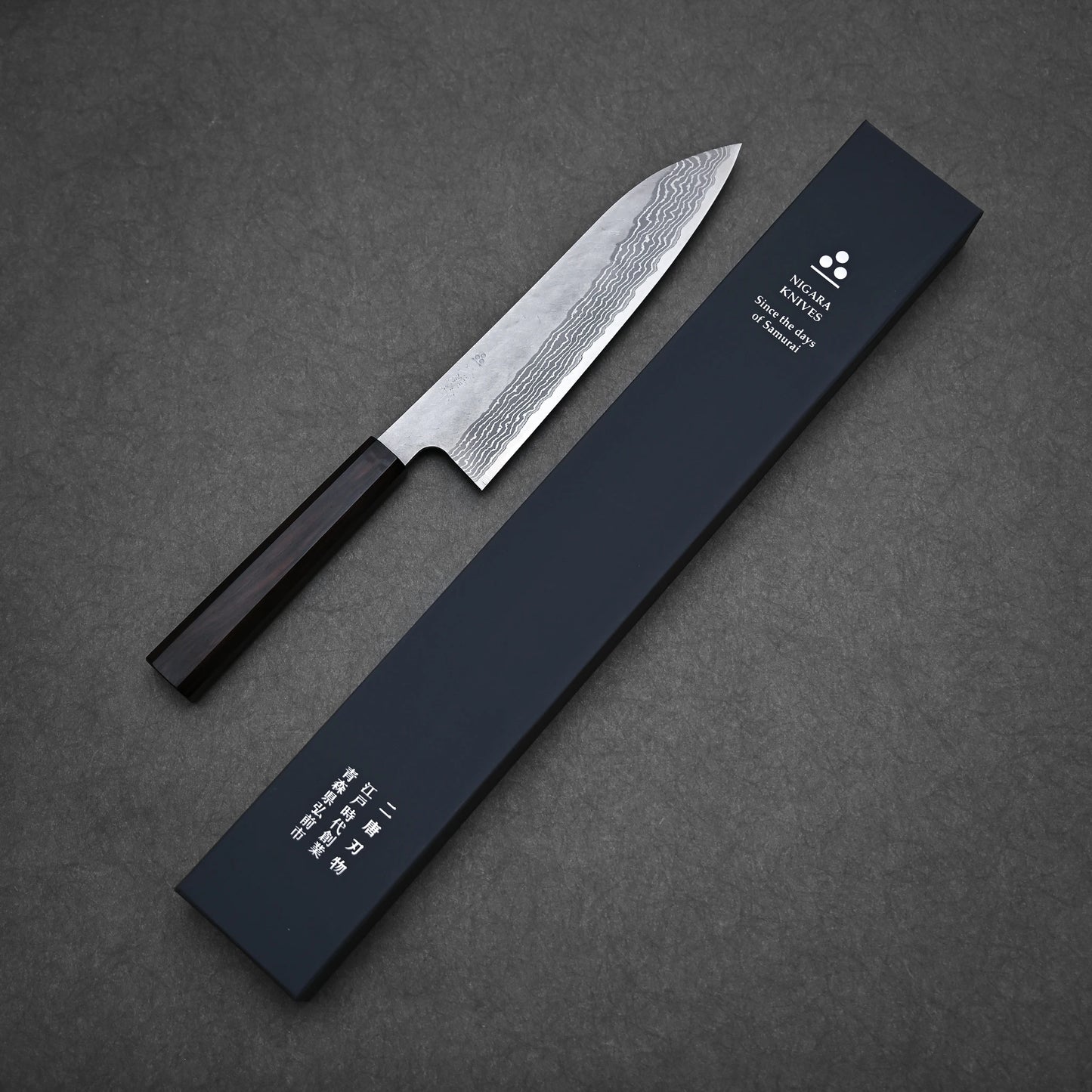 A Nigara Hamono tsuchime damascus VG10 gyuto 210mm with a dark handle is displayed next to a long, dark blue box with white Japanese writing and branding on a gray textured surface.