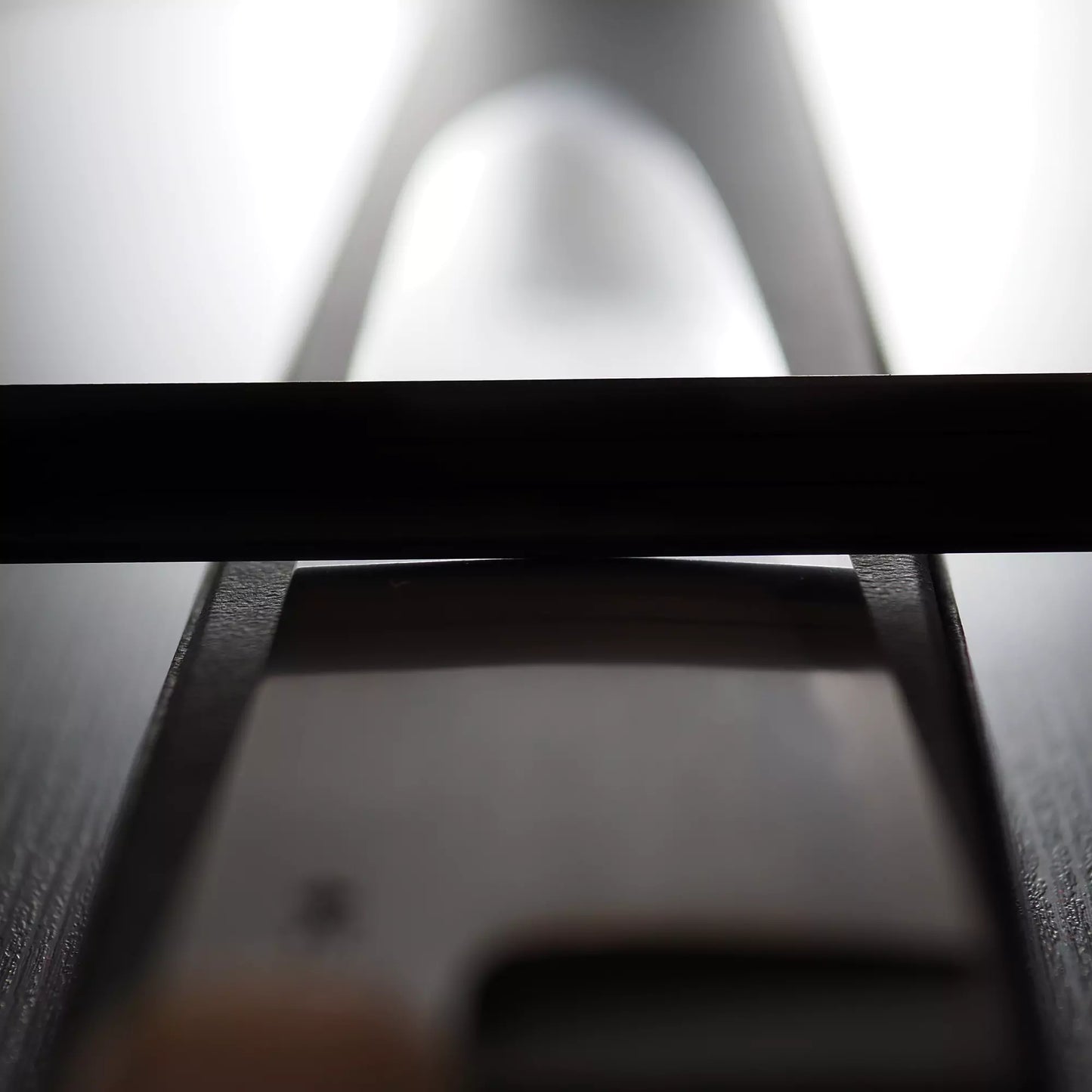 Close-up of the Satoshi Nakagawa bokashi shirogami#1 tall gyuto 240mm, highlighting its blade and handle beneath a black object—possibly a cutting board—on a dark surface with bright backlighting.