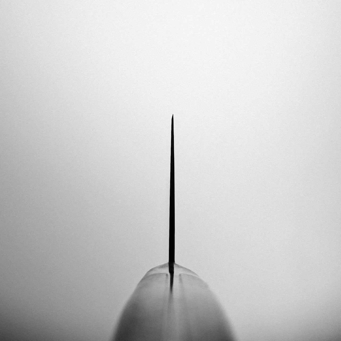 A black and white photo featuring the tip of the Misono Molybdenum santoku 180mm knife, centered and rising vertically against a blurred, light background.