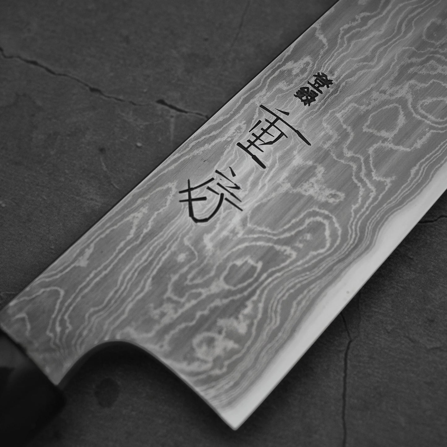 A close-up of a knife's blade, showcasing its unique Damascus steel pattern and distinct kanji character engravings. It rests on a dark background.
