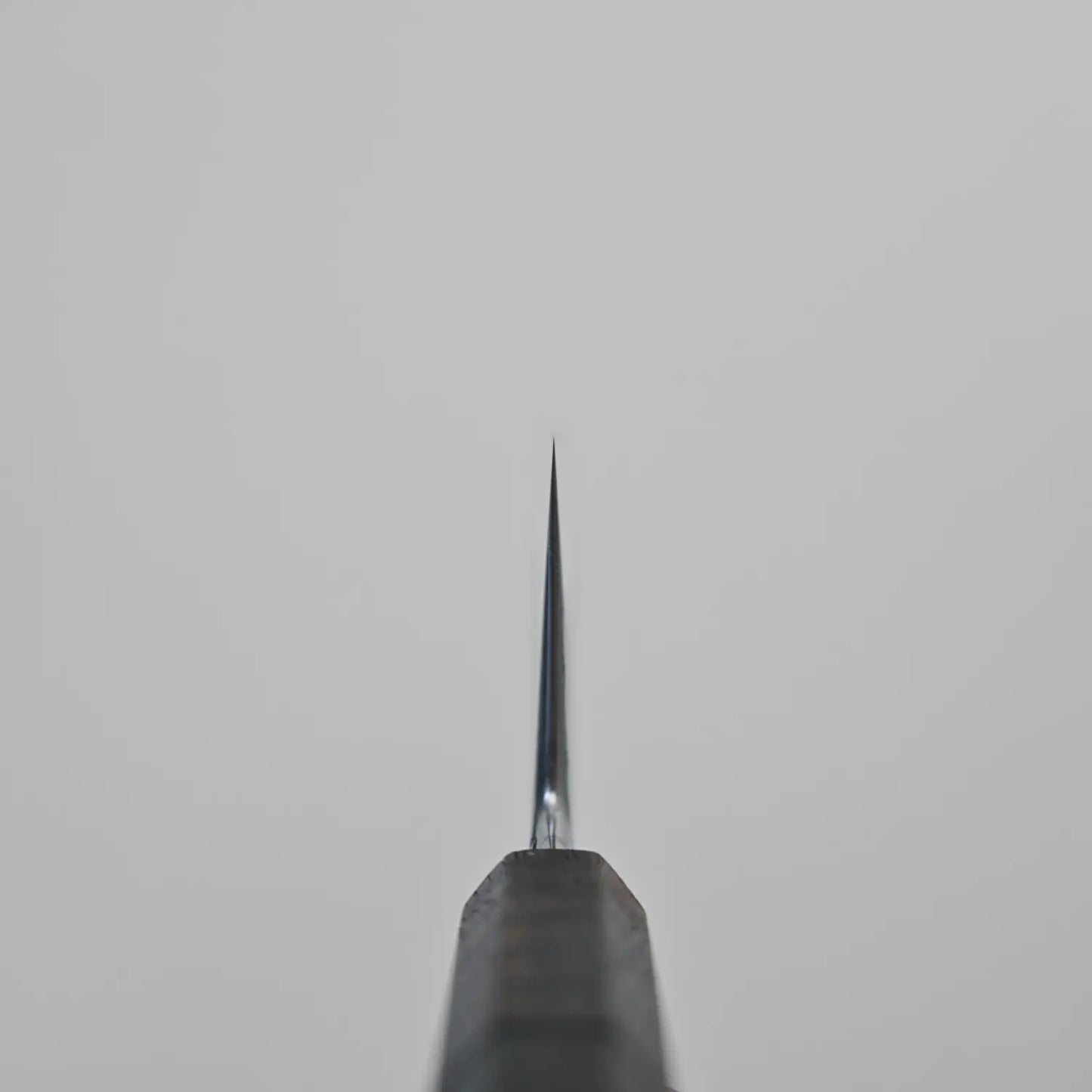 Close-up of the sharp tip of a Krichbaum kurouchi wrought iron clad Apex Ultra gyuto 215x55 blade, photographed from above and centered against a plain light grey background.