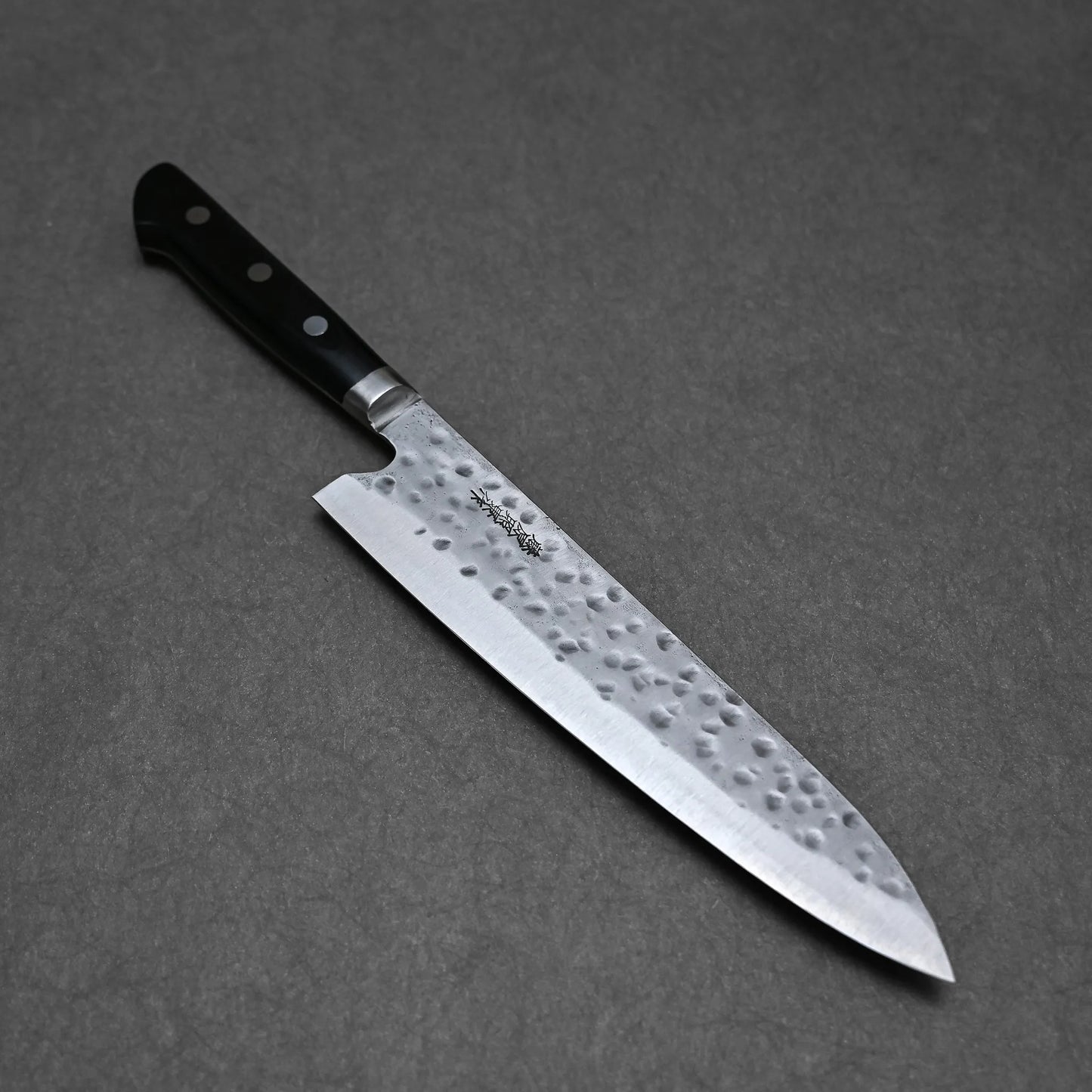 The Teruyasu Fujiwara Maboroshi shirogami#1 gyuto 240mm, featuring a hammered stainless steel blade and a triple-riveted black handle, is shown resting on a dark gray textured surface.