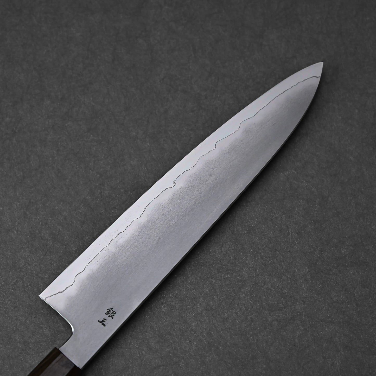 A full view of the reverse side of a gyuto knife blade. The ginsan stainless steel blade features a kasumi finish with a clear, wavy border extending along its length and tapering to a pointed tip. Kanji engravings are visible near the heel of the blade.