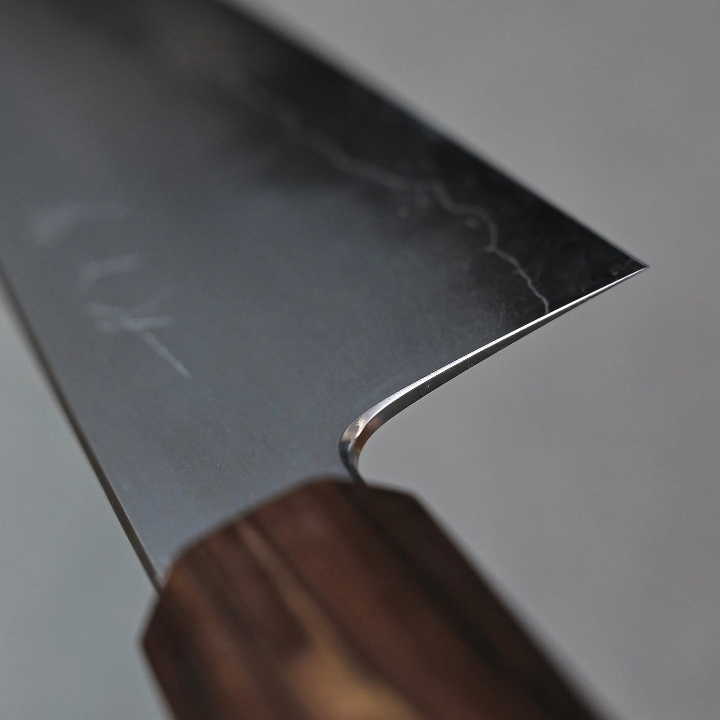 A close-up, angled view of the choil of a gyuto knife, where the ginsan stainless steel blade smoothly transitions into the mono octagonal ebony handle. The polished cutting edge is visible curving away from the choil towards the right.