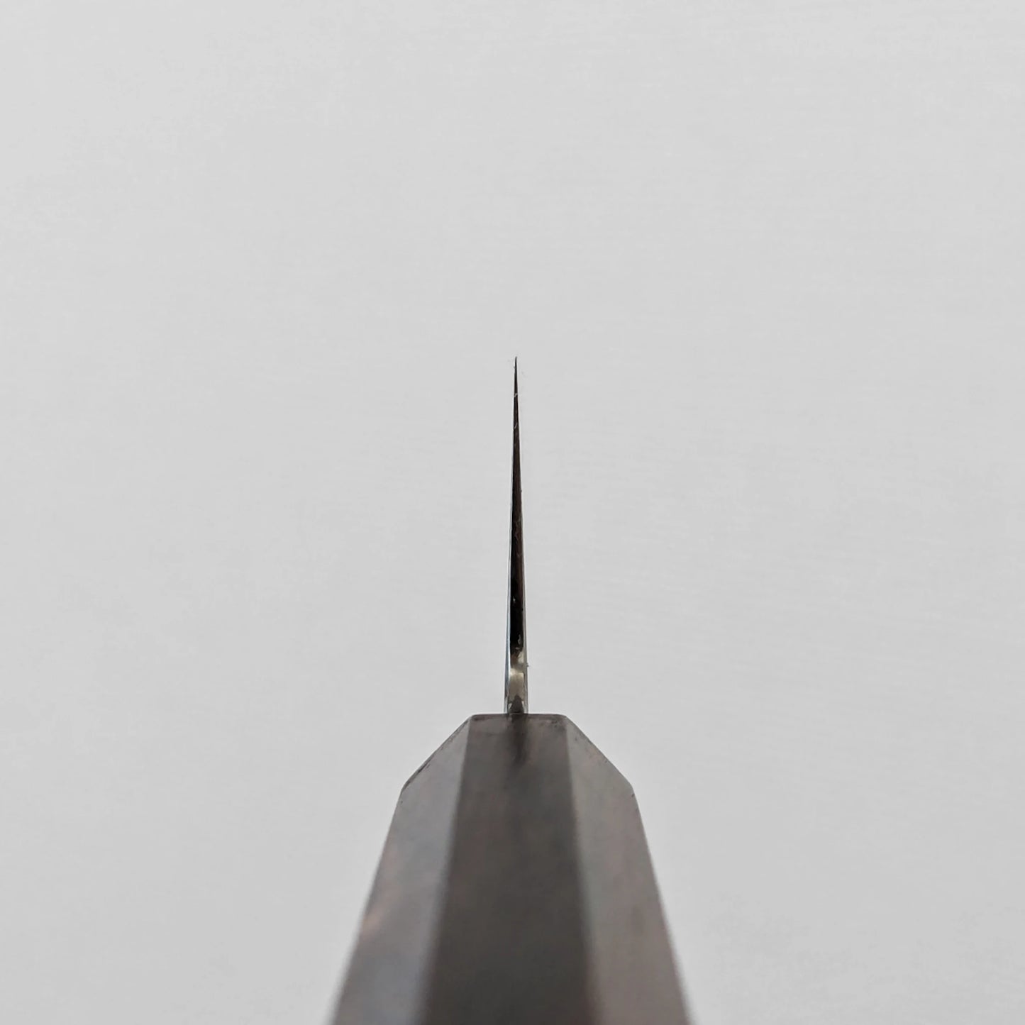 A vertical close-up shot of the choil of a gyuto knife, seen from directly below. The polished ginsan stainless steel blade tapers sharply upwards to a fine point, emerging from the dark mono octagonal ebony handle, which occupies the lower portion of the frame.