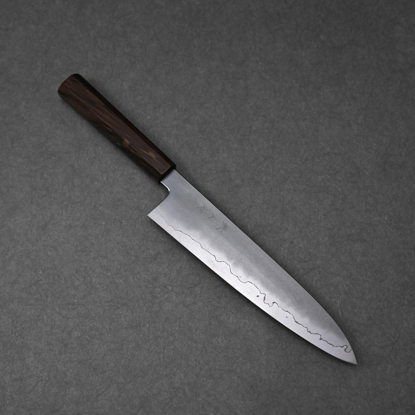 A full, overhead view of a gyuto knife, positioned diagonally on a textured grey surface. The ginsan stainless steel blade displays a kasumi finish with a prominent wavy border and subtle kanji engravings on its upper surface. The knife is fitted with a dark, mono octagonal ebony handle.