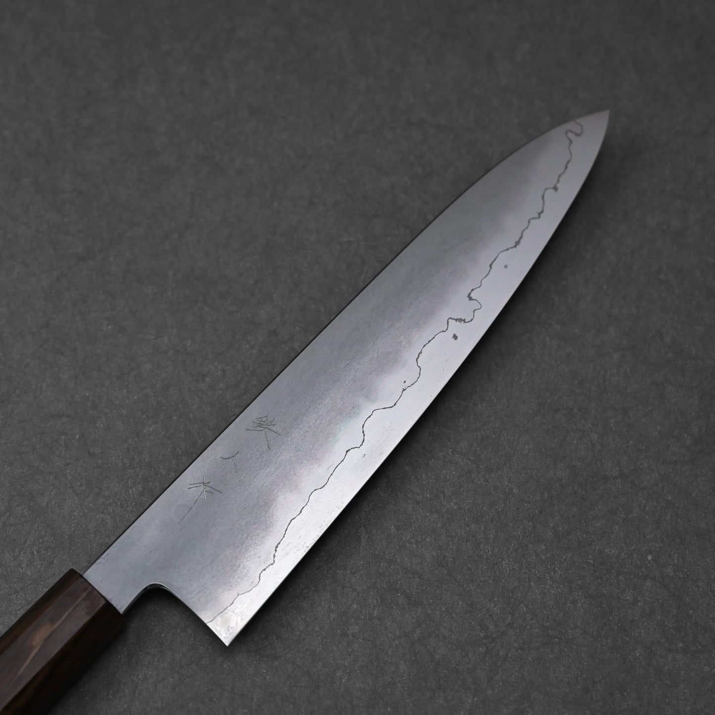 A close-up of a gyuto knife blade, made from ginsan stainless steel. The kasumi finish is clearly visible, showing a frosted upper portion with subtle kanji engravings, and a polished lower edge following a distinct wavy line. The blade tapers towards a rounded tip.