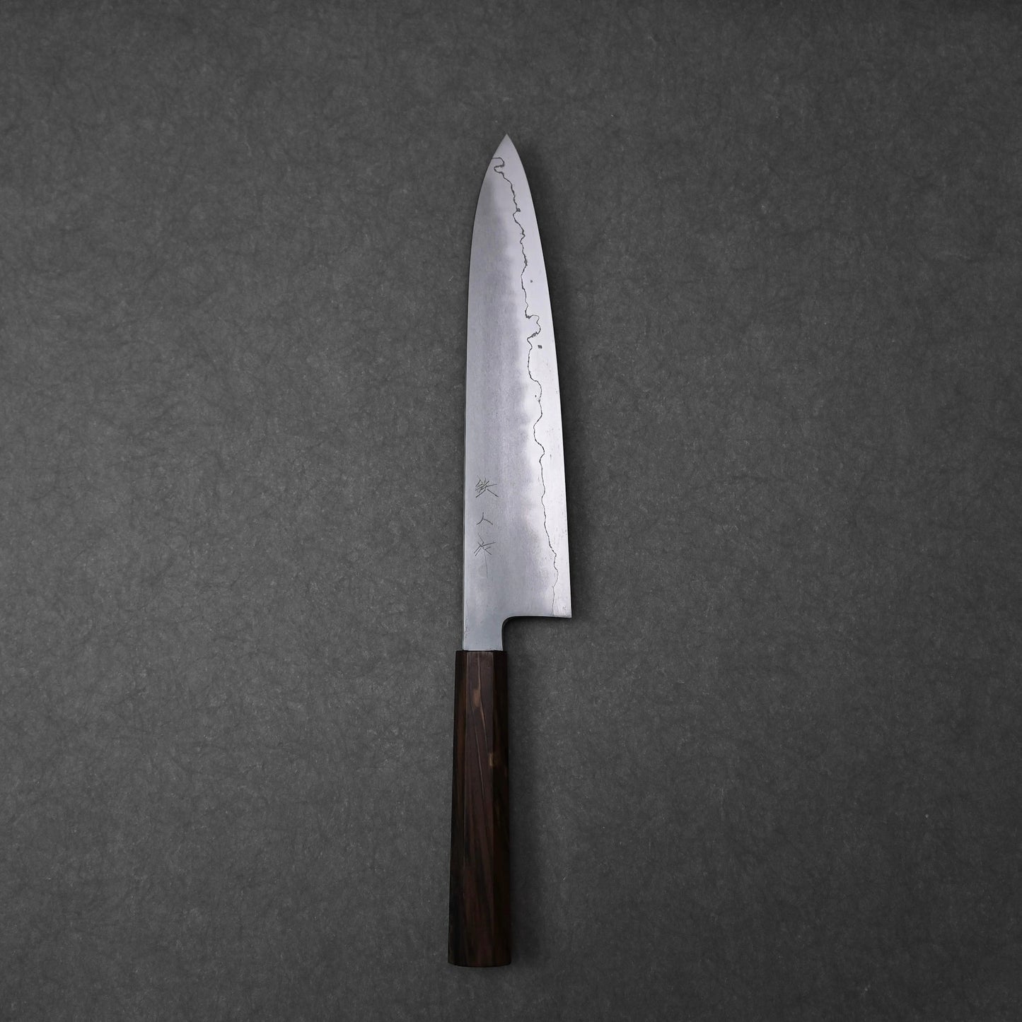 A full, overhead view of a gyuto knife, positioned vertically on a dark grey surface. The ginsan stainless steel blade features a kasumi finish with a wavy border and kanji engravings on the upper surface. The knife has a dark, mono octagonal ebony handle at the bottom.