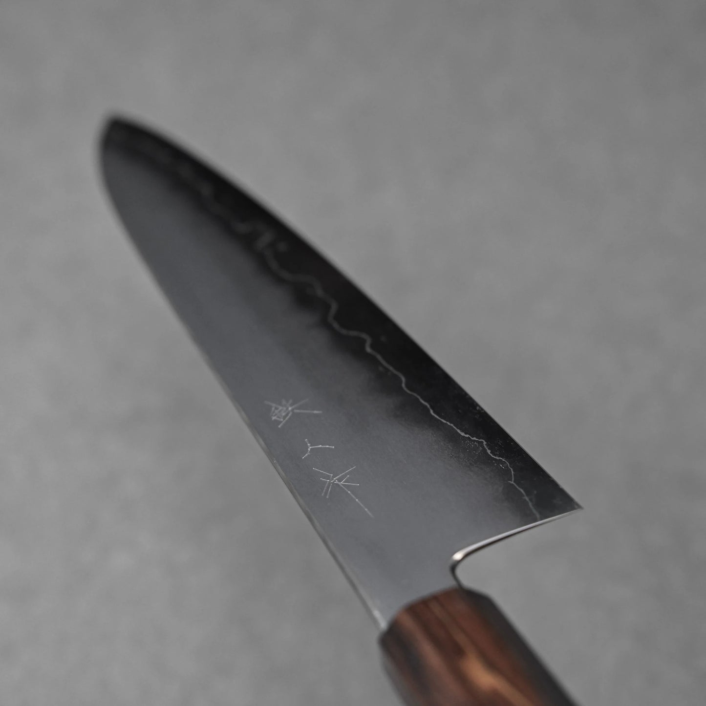 A close-up, angled view of a gyuto knife blade. The ginsan stainless steel blade shows a distinct kasumi finish with a darker, frosted upper section and a wavy polished edge. Subtle kanji characters are engraved on the upper part of the blade, which tapers towards a rounded tip that is out of focus. Part of the dark wooden handle is visible at the bottom.