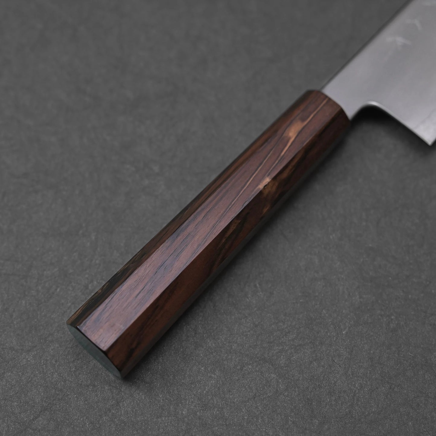 A close-up view of the mono octagonal ebony handle of a gyuto knife. The dark wood exhibits rich, varied brown tones and a visible grain pattern, with the eight facets of its shape clearly defined along its length.