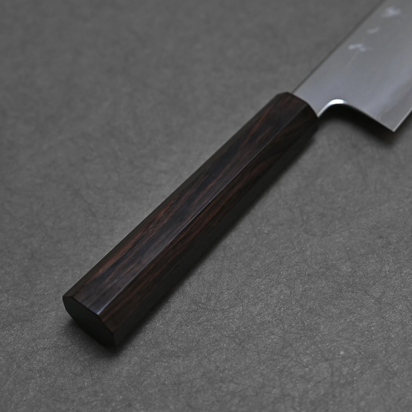 A close-up view of the dark, octagonal ebony handle of a kiritsuke gyuto knife. The rich grain of the wood is subtly visible, and the facets of its eight-sided shape are clearly defined.