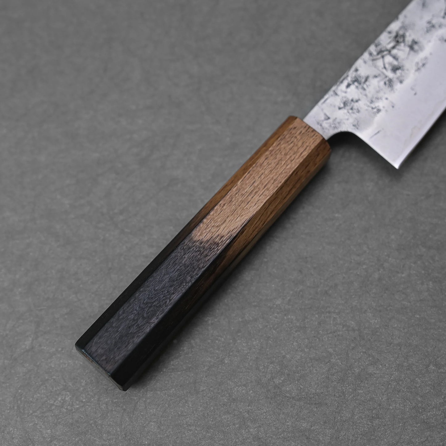 A close-up of the Tsunehisa nashiji SLD bunka 170mm knife, featuring an SLD steel blade and a wooden handle with three wood tones, resting on a textured gray surface.