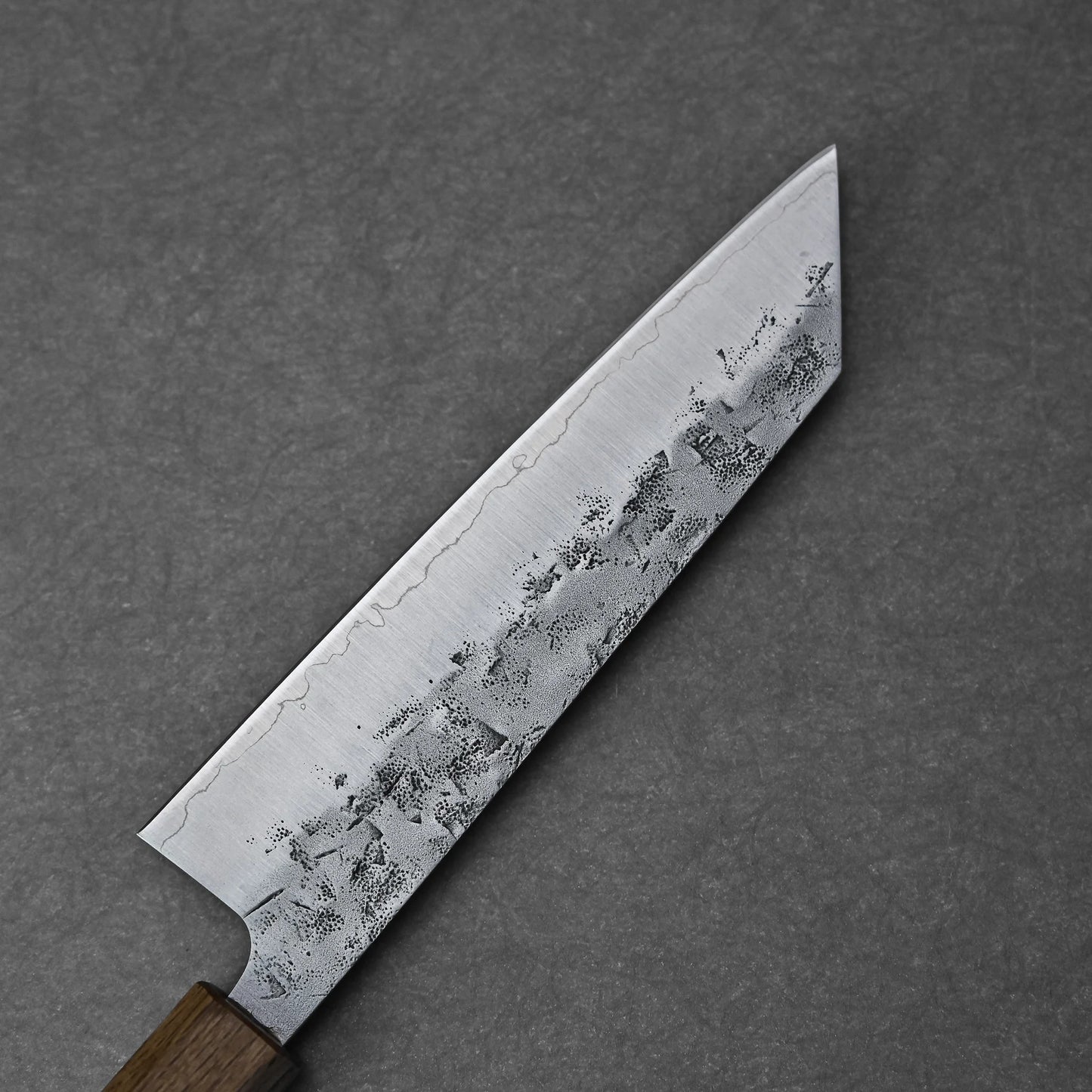 A close-up of the Tsunehisa nashiji SLD bunka 170mm knife, featuring a wooden handle and patterned SLD steel blade, resting on a dark gray textured surface.