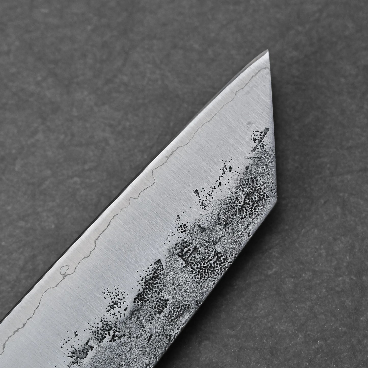 Close-up of the Tsunehisa nashiji SLD bunka 170mm blade tip, highlighting intricate patterns and a wavy hamon line on SLD steel against a dark textured background.