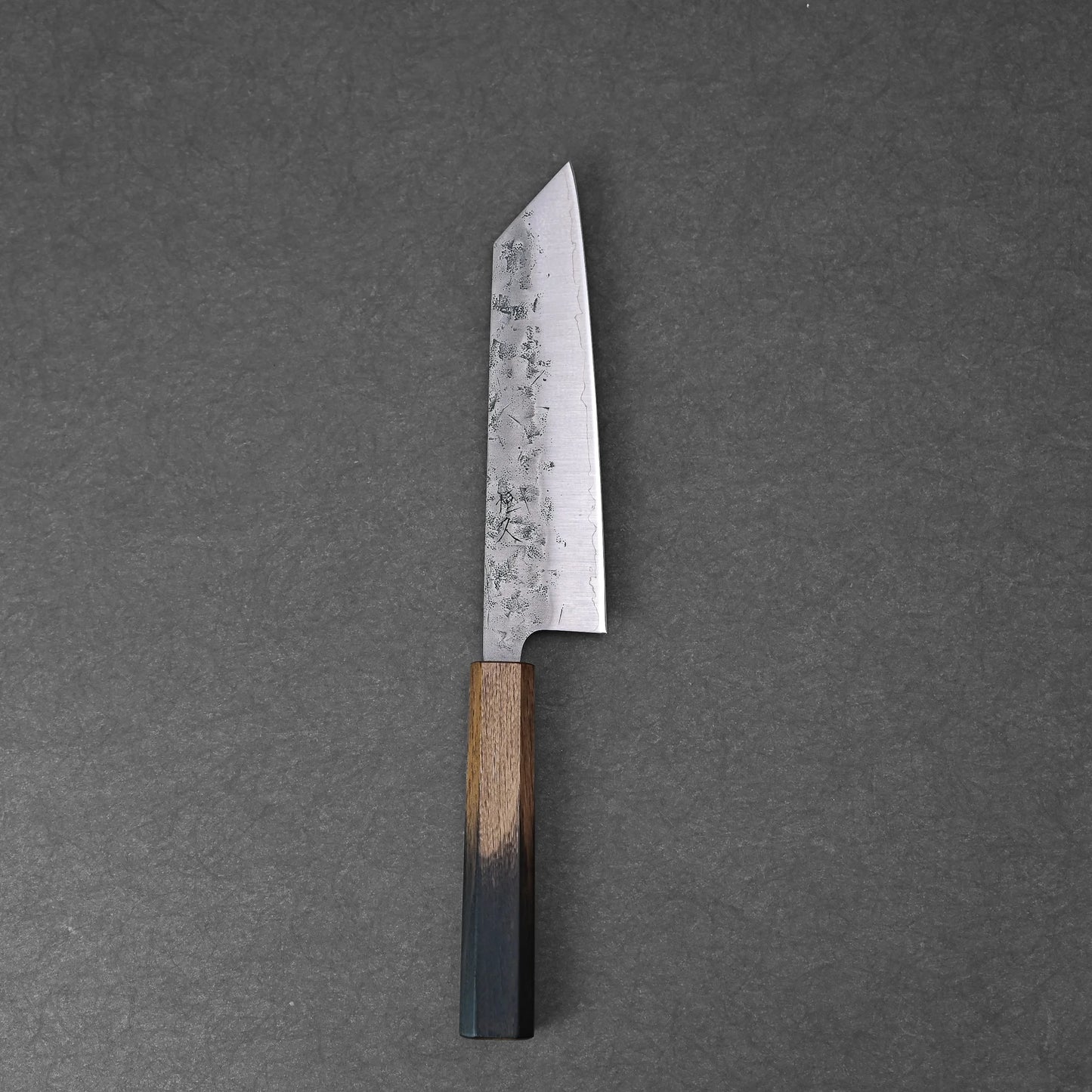 The Tsunehisa nashiji SLD bunka 170mm, featuring a straight-edged hammered SLD steel blade and a wooden handle, rests on a dark grey textured surface.