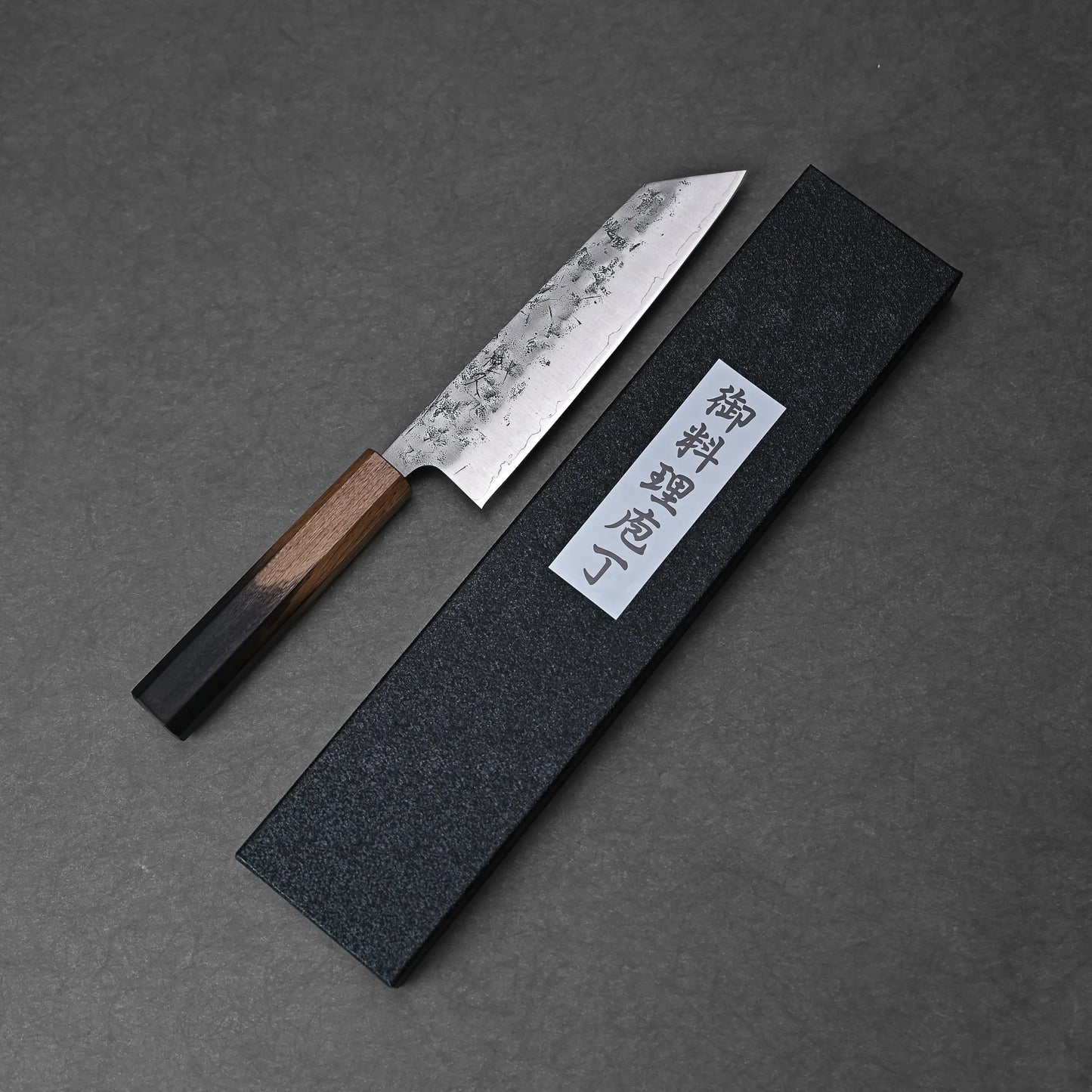 The Tsunehisa nashiji SLD bunka 170mm—a Japanese kitchen knife with a wooden handle—rests beside a black textured box with a white Japanese label, all arranged on a dark gray surface.