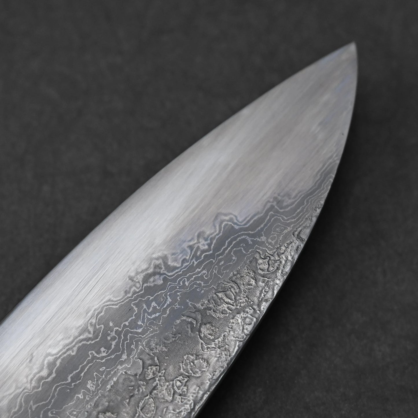 Reverse side of a Japanese knife tip featuring damascus steel with layered wave patterns and subtle forging textures; photographed close-up to highlight material contrast and pointed edge detail.