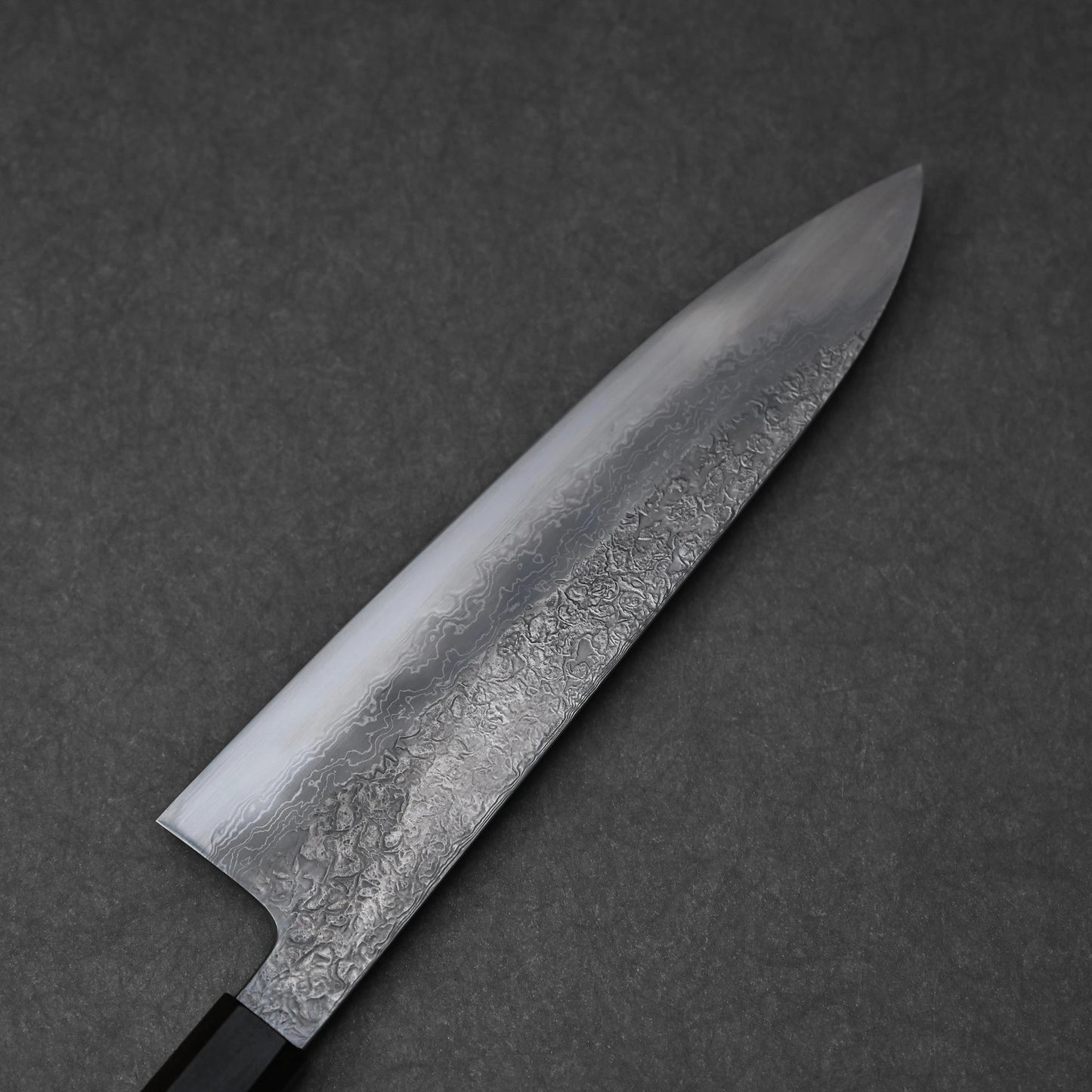 Reverse side of a Japanese knife blade featuring damascus steel patterns with layered waves and subtle forging marks; photographed close-up to highlight craftsmanship and steel texture.
