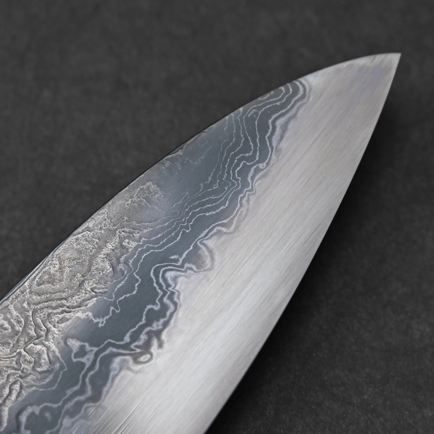 Close-up of the knife tip featuring damascus steel patterns with layered waves and subtle forging marks; photographed to emphasize the pointed edge and material intricacy.