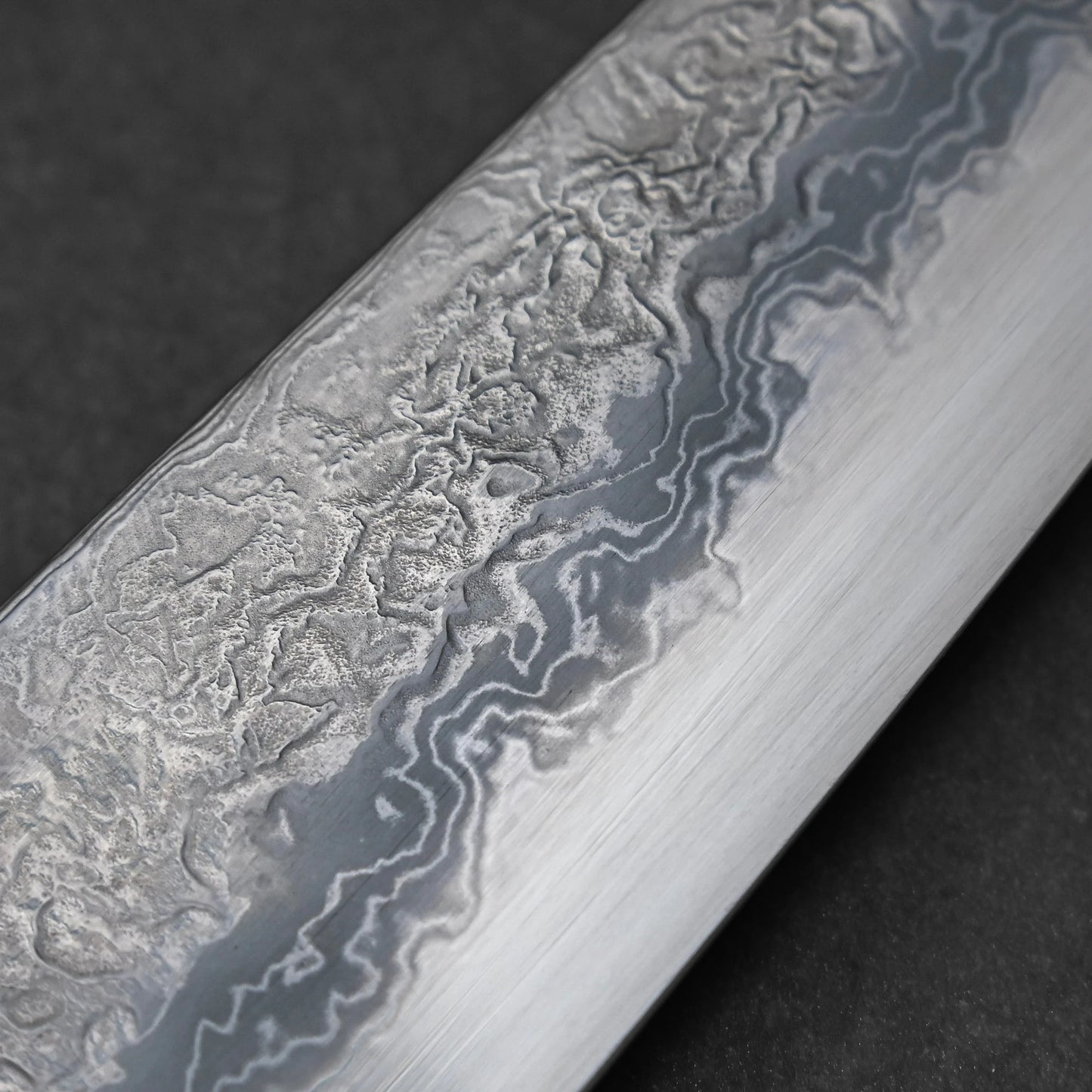 Midsection of a Japanese knife blade featuring damascus steel patterns with layered waves and subtle forging marks; photographed close-up to highlight the steel's texture complexity and craftsmanship detail.