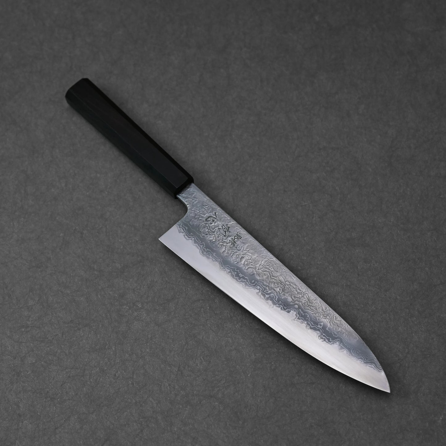 Japanese chef’s knife featuring damascus steel wave patterns and vertically engraved kanji characters near the handle; photographed at a standard angle to highlight the full blade and engraving placement.
