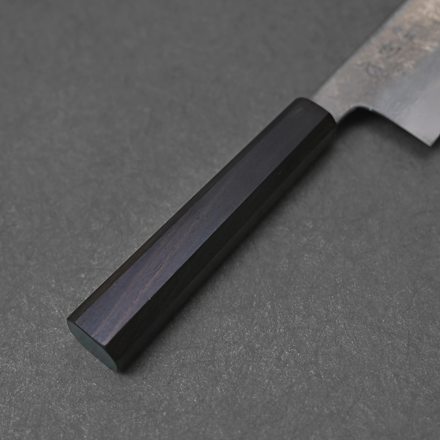 Handle of a Japanese knife featuring a dark octagonal shape with a smooth polished finish; photographed close-up against a textured dark surface to emphasize material quality and craftsmanship detail.