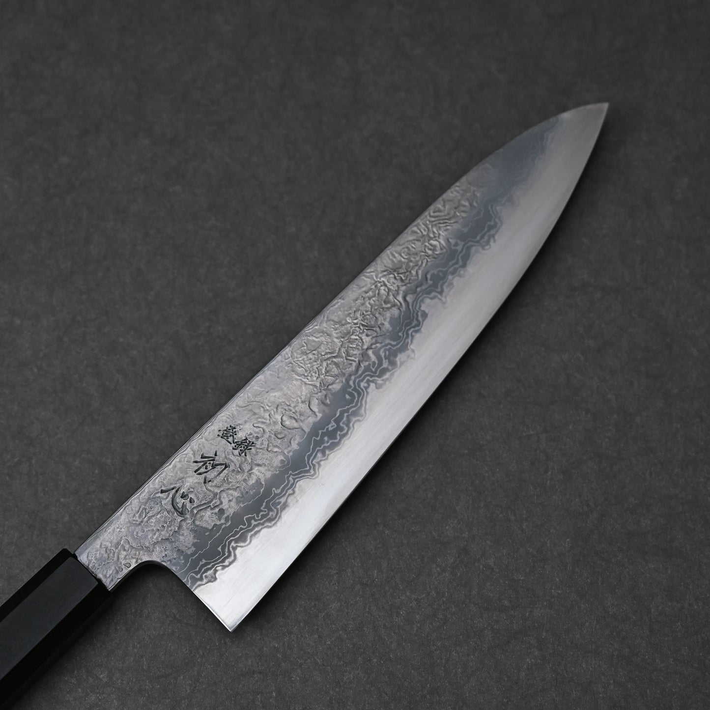 Japanese chef’s knife with damascus steel blade featuring layered forge patterns and vertically engraved kanji near the handle; paired with a black octagonal handle and photographed against a dark textured background.