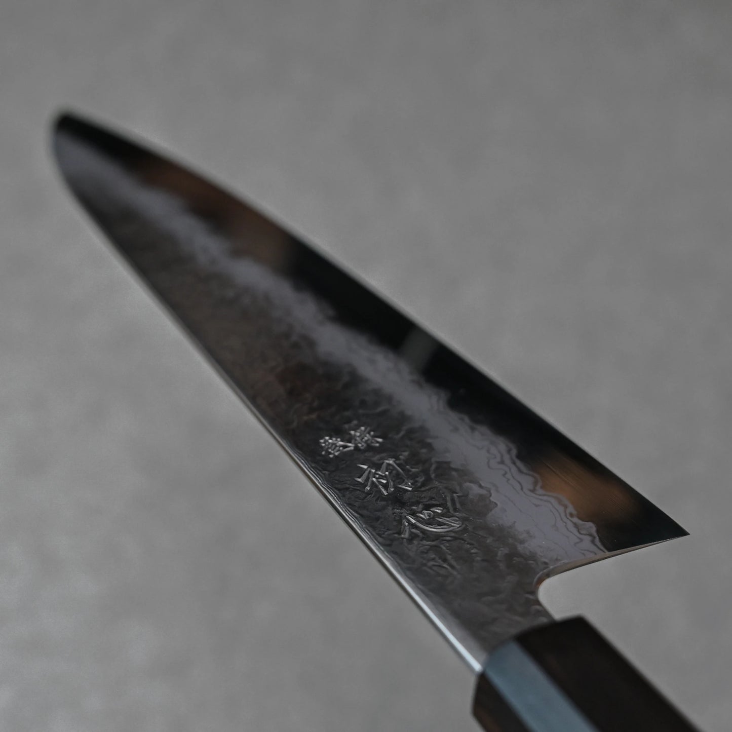Close-up of a Japanese knife blade featuring damascus steel patterns with layered wave textures and vertically engraved kanji near the handle; photographed to highlight fine craftsmanship and artistic detail.