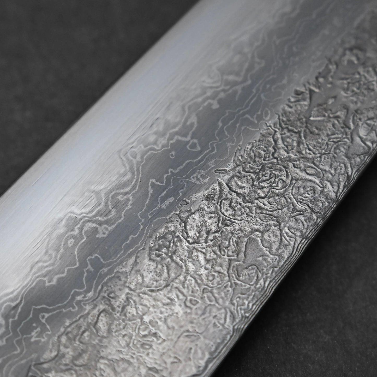 Reverse side midsection of a Japanese knife blade featuring damascus steel construction with layered wave patterns and subtle forging textures; photographed close-up to showcase steel intricacy and material contrast.
