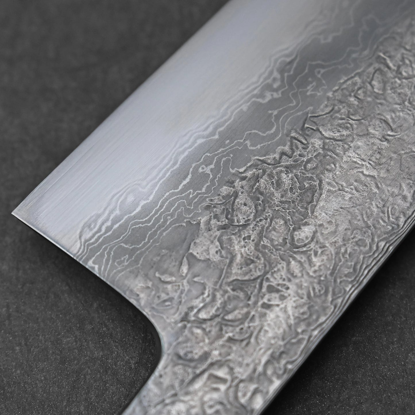 Japanese knife blade featuring damascus steel construction with layered wave patterns and subtle forging marks across the midsection; paired with a black octagonal handle and photographed diagonally against a neutral background for balanced contrast.