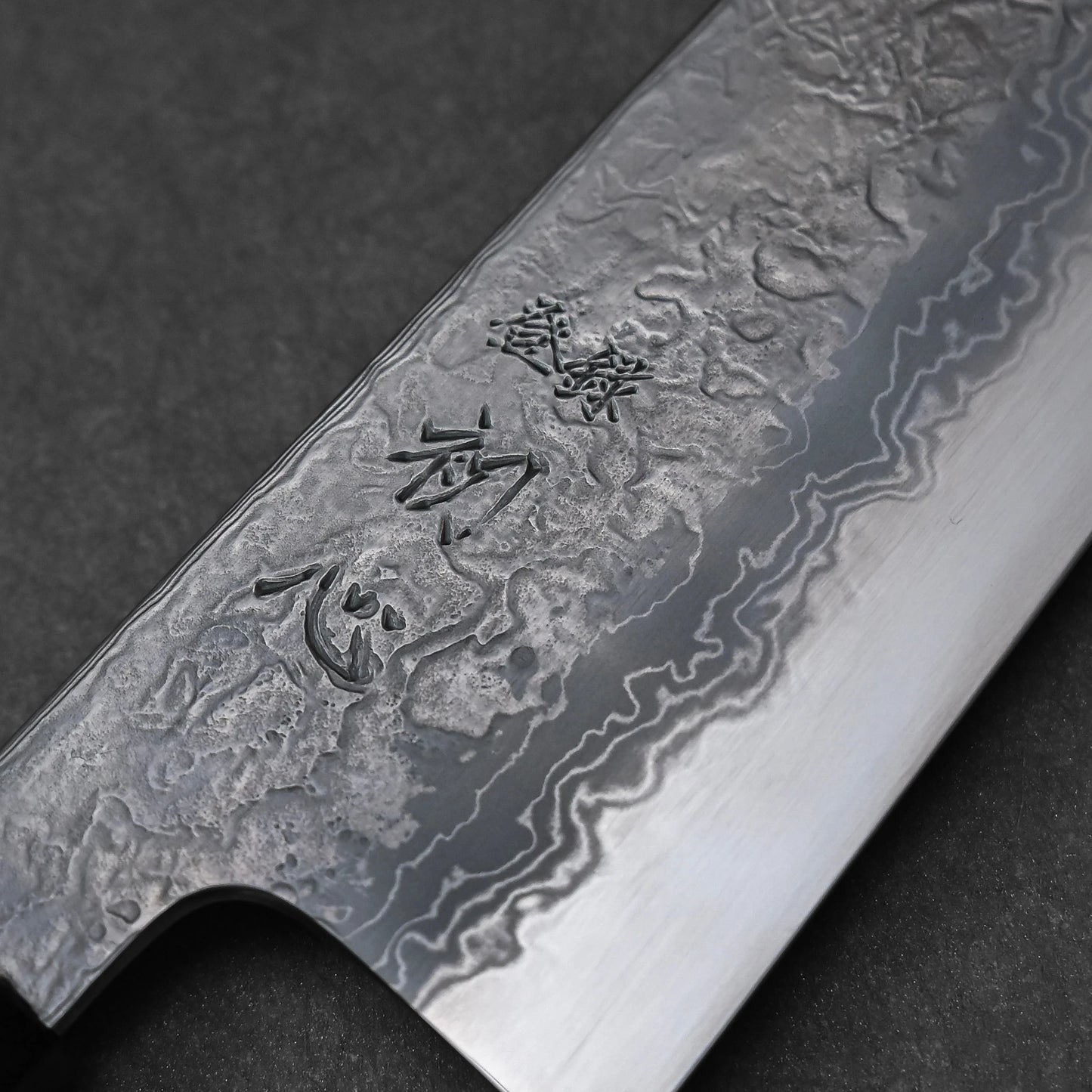 Close-up of a Japanese knife blade featuring damascus steel patterns with vertically engraved kanji characters near the spine; photographed to emphasize the blade’s layered texture and traditional artistry.