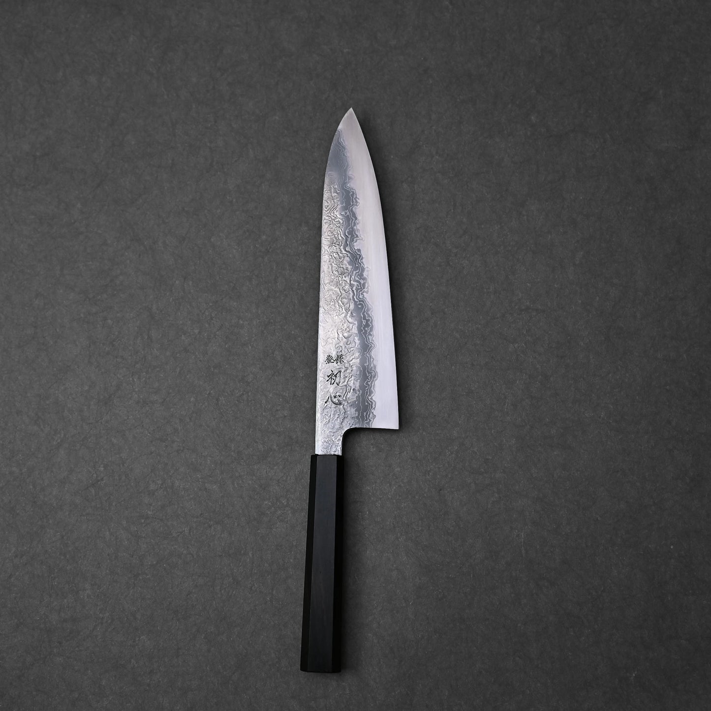 Japanese chef’s knife with damascus steel blade showcasing layered wave patterns and vertically engraved kanji near the handle; paired with a black octagonal handle and photographed flat against a dark textured surface to highlight craftsmanship and design detail.