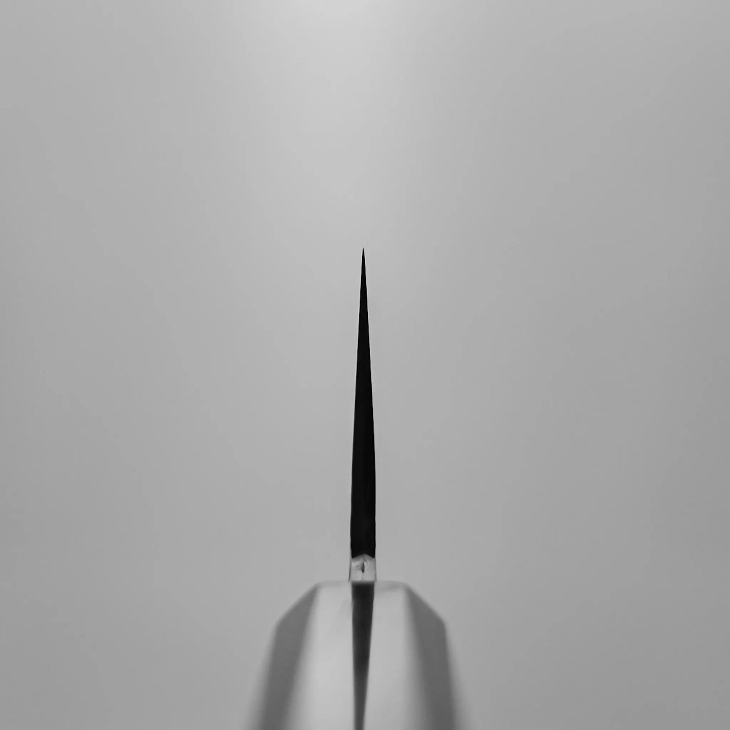 Close-up of the Yoshihide Masuda aogami#1 damascus santoku 180mm’s sharp edge, shown vertically with its point facing upward and centered against a plain light gray background.