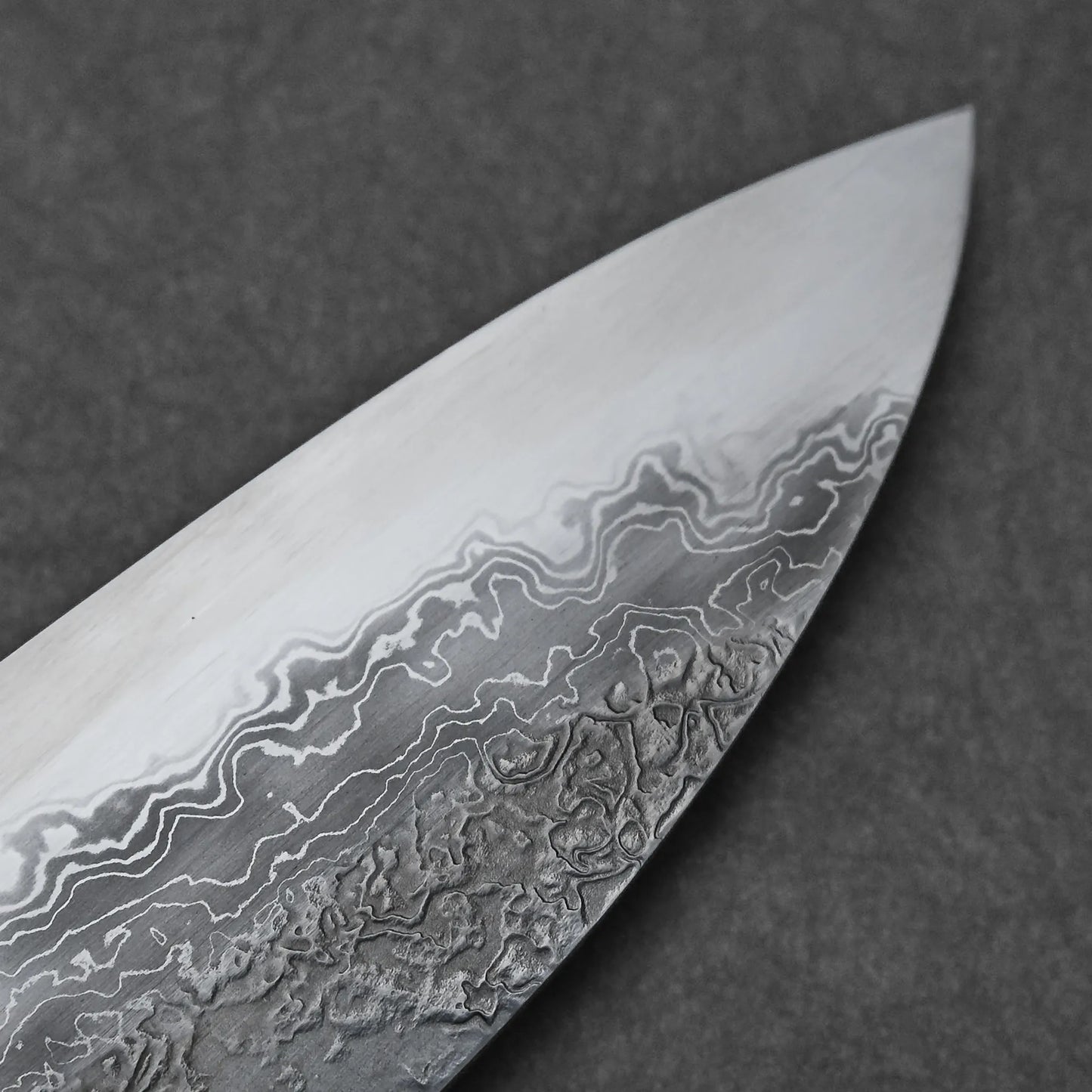 Close-up of the Yoshihide Masuda aogami#1 damascus santoku 180mm blade, highlighting its damascus finish with wavy, intricate lines and textures; set against a plain dark gray background.
