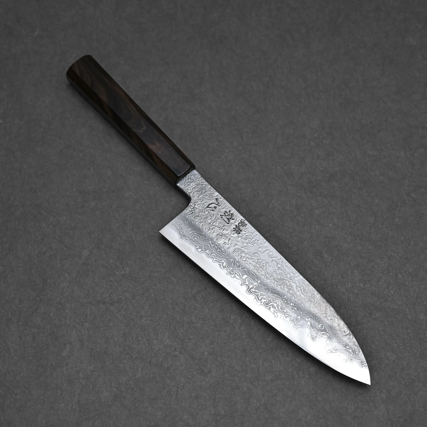 The Yoshihide Masuda Aogami#1 Damascus Santoku 180mm, featuring a dark wooden handle and handcrafted Damascus-finish blade, rests on a dark gray surface.