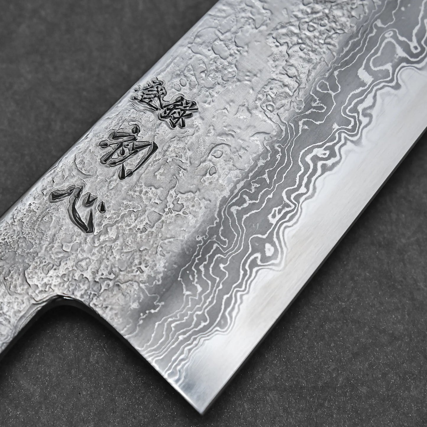 Close-up of the Yoshihide Masuda aogami#1 damascus santoku 180mm blade, featuring a striking Damascus finish and engraved Japanese characters, showcasing its layered metal texture against a dark background.
