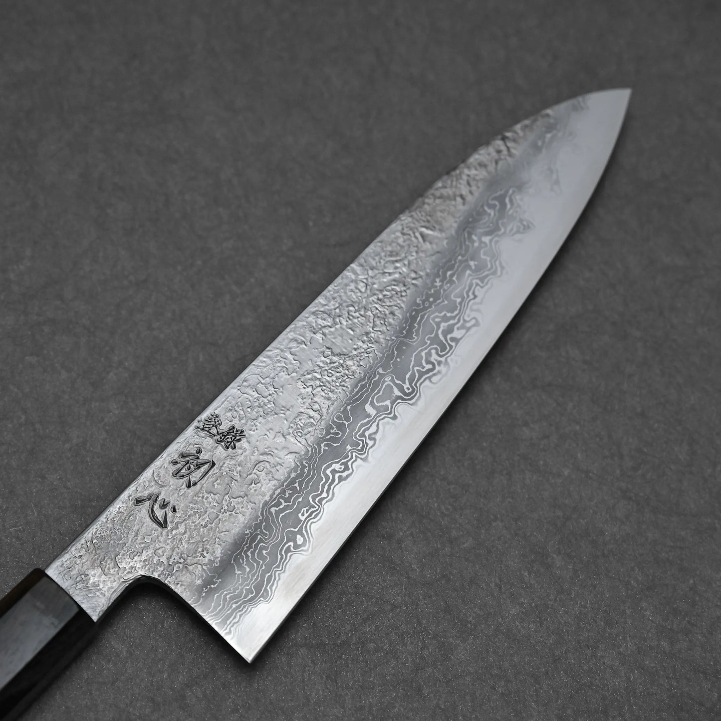 A close-up of the Yoshihide Masuda aogami#1 damascus santoku 180mm shows its wavy-patterned damascus blade with engraved Japanese characters, set against a plain gray background.