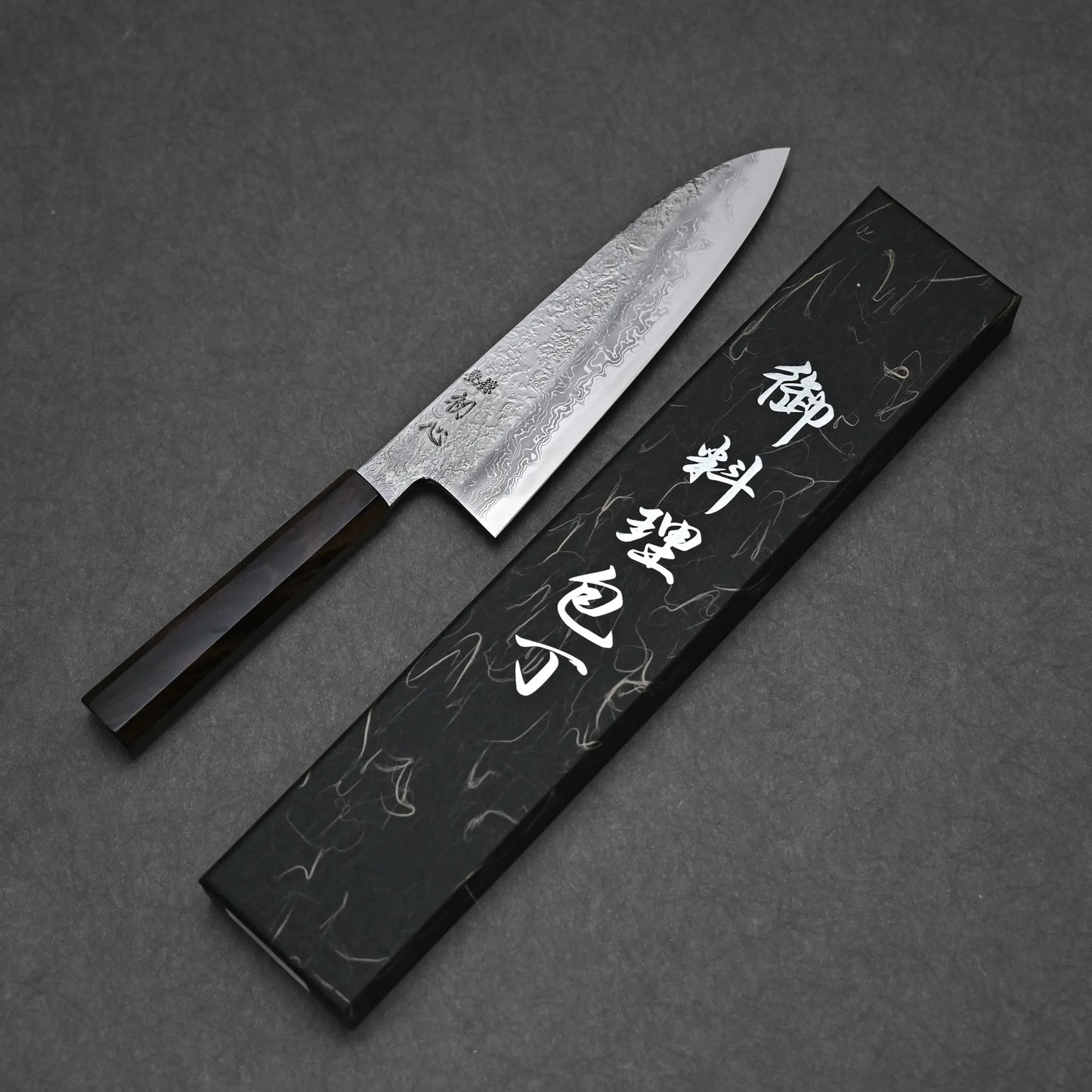The Yoshihide Masuda aogami#1 damascus santoku 180mm, featuring a damascus finish and dark wooden handle, rests beside a black box with white Japanese lettering on a dark surface.