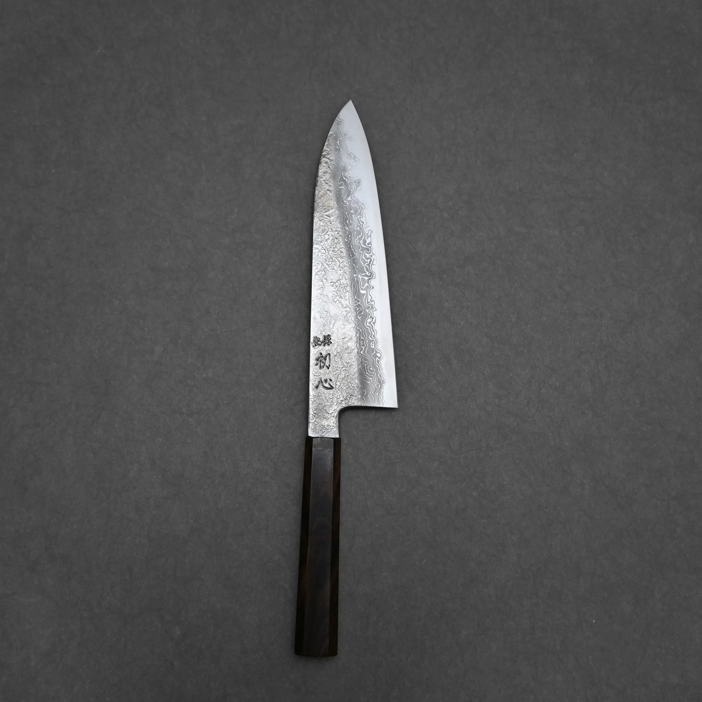 The Yoshihide Masuda aogami#1 damascus santoku 180mm, featuring a damascus blade and dark wooden handle, rests on a plain dark gray surface.