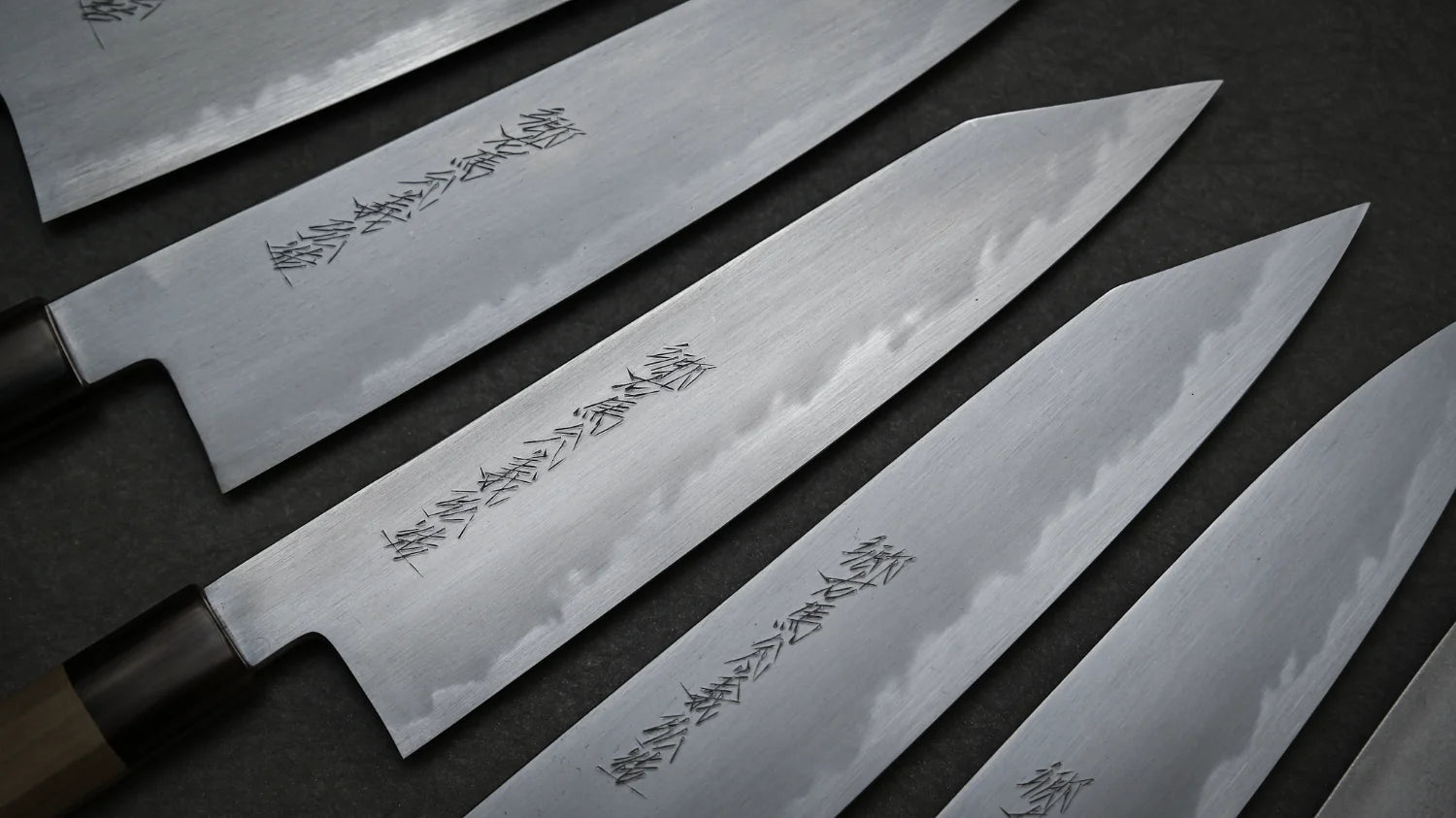 Japanese kitchen knives with carbon steel blades arranged diagonally on a dark surface. The knives are Yoshikazu Tanaka shirogami#1 gyuto knives with engraved Japanese characters.