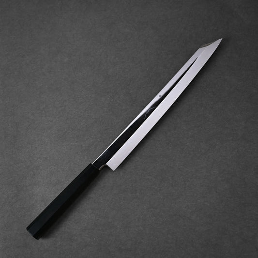 A Yoshikazu Ikeda shirogami#1 honyaki kiritsuke yanagiba 330mm Japanese kitchen knife with a straight black handle and a long, slender blade rests on a dark gray surface.