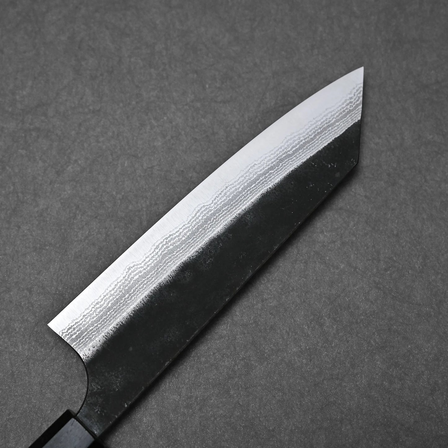 A Yoshimi Kato kurouchi damascus AS bunka 170mm knife with a dark handle and patterned Aogami Super steel blade is shown close-up on a gray textured surface.