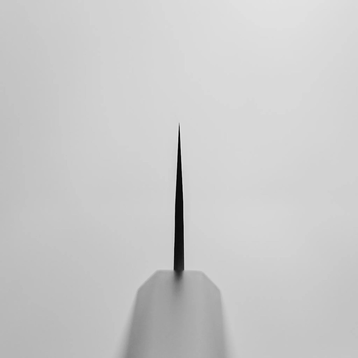 A close-up, top-down view of a Yoshimi Kato kurouchi damascus AS bunka 170mm’s pointed tip on a plain light gray background. The out-of-focus handle sits at the bottom while the Aogami Super steel blade tapers to a fine point in the center.