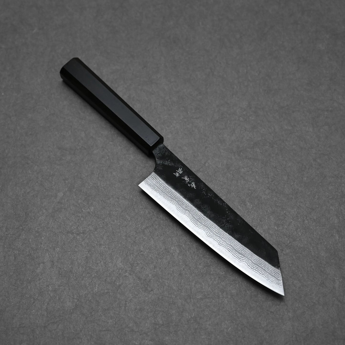 A Yoshimi Kato kurouchi damascus AS bunka 170mm knife with a dark handle and wide patterned blade rests on a flat, dark gray surface. Subtle markings are visible near the handle on its Aogami Super steel blade.