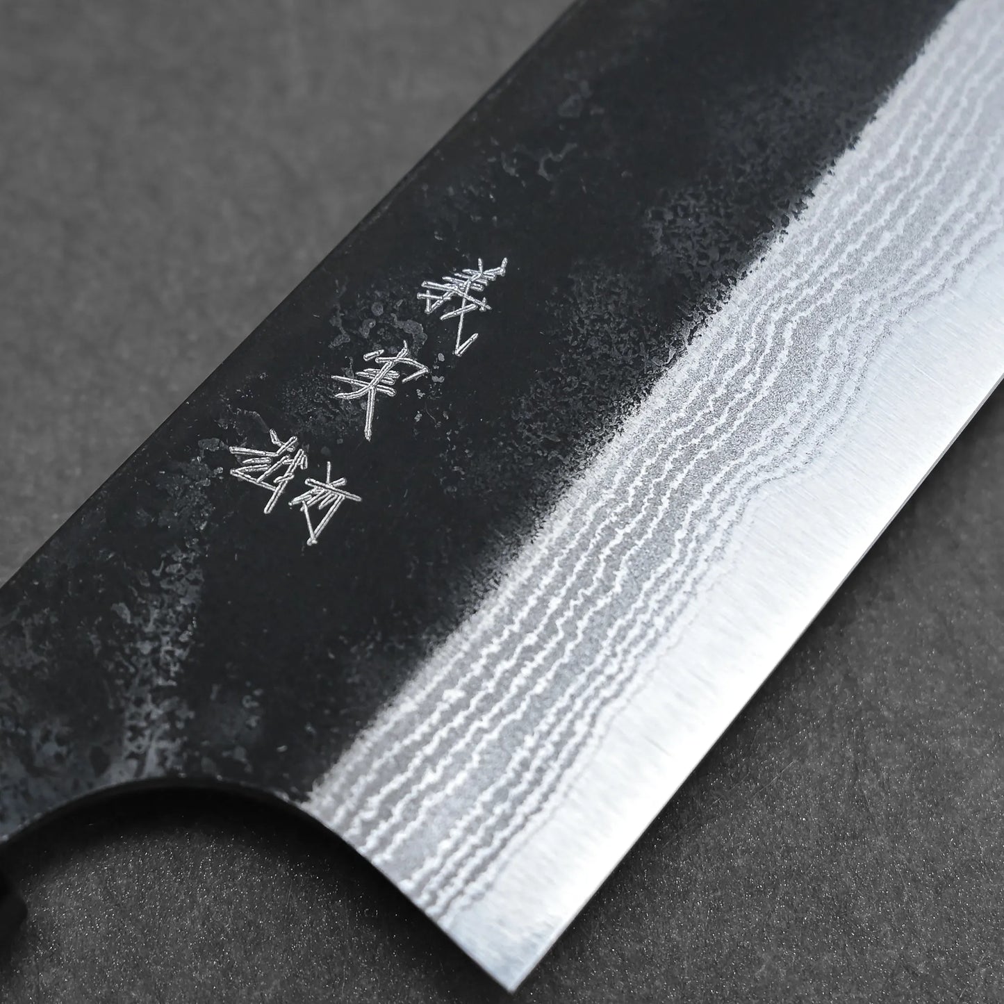 Close-up of the Yoshimi Kato kurouchi damascus AS bunka 170mm blade, featuring wavy damascus patterns and engraved Japanese characters on the dark upper section. Made from Aogami Super steel for outstanding performance against a textured background.