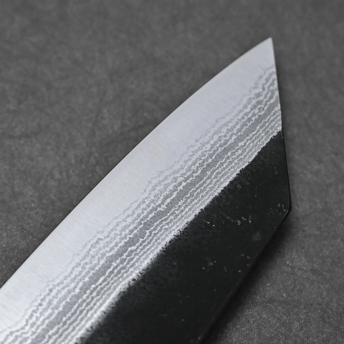 Close-up of the tip of a Yoshimi Kato kurouchi damascus AS bunka 170mm knife blade, showcasing its unique wavy Damascus patterns against a dark background.